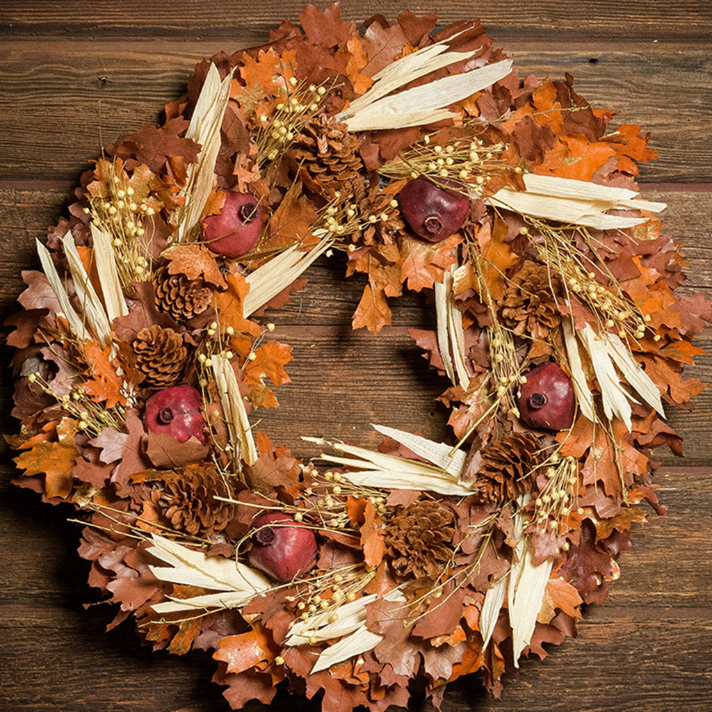 A close-up of a dried wreath made of cafe colored preserved oak leaves, natural corn husk, amber oak leaves with hints of gold-marbled accents, natural pinecones, and natural ruby pomegranates hung on a dark wood background.