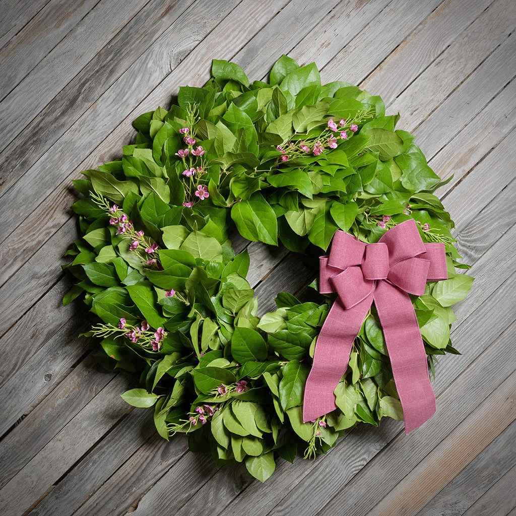Green leaf wreath with pink flowers and a bow on a wooden surface