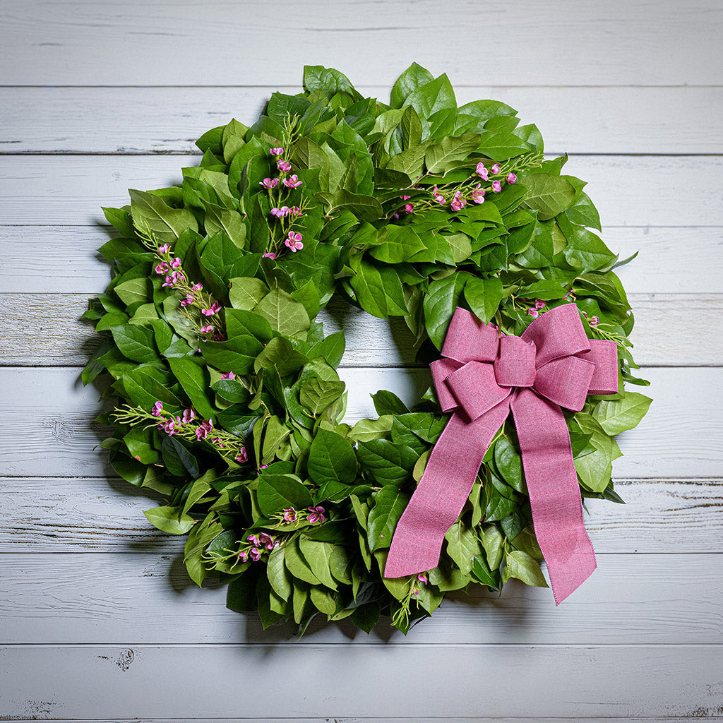 Green leafy wreath with pink flowers and a pink bow on a white wooden background