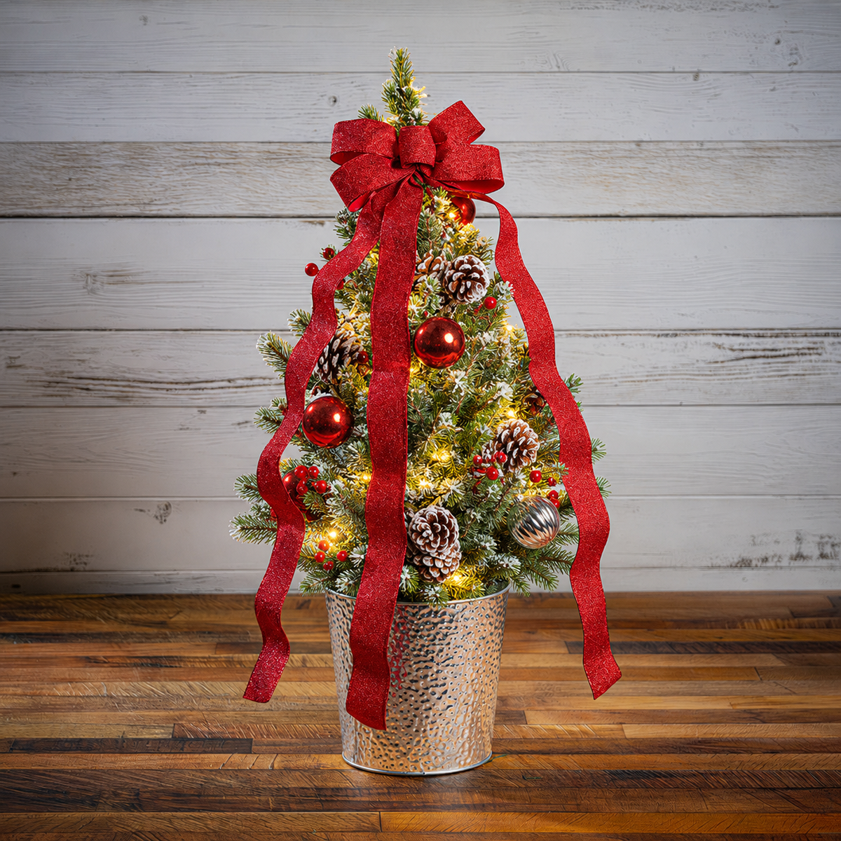 A small artificial Christmas tree in a hammered silver bucket planter, decorated with a large red ribbon bow, red ornaments, pinecones, and warm white string lights, set against a rustic white wood plank wall and dark wood floor.