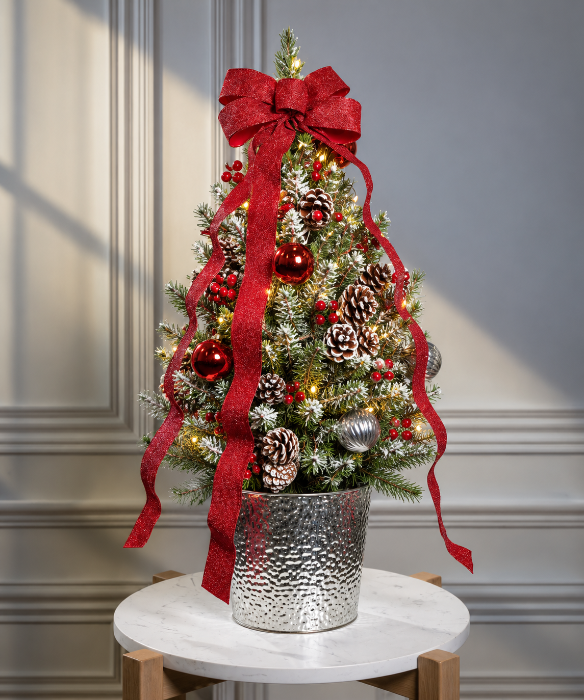 A small artificial Christmas tree with white snow-like dusting, red ornaments, pinecones, warm white string lights, and a large red ribbon bow, displayed in a textured silver pot on a round white table against a gray paneled wall.