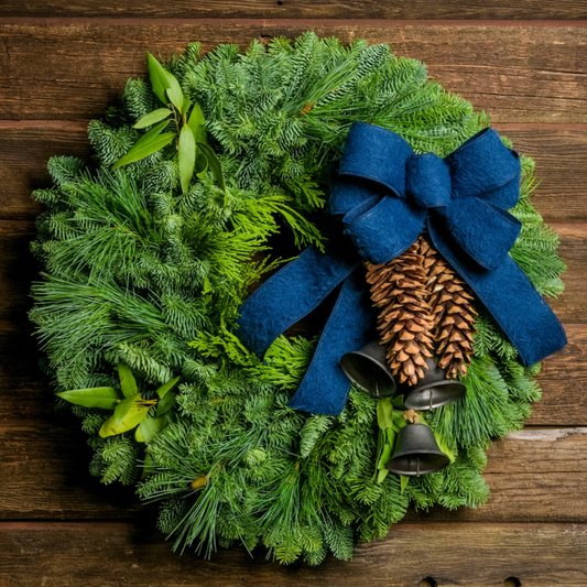 Holiday wreath made of noble fir, incense cedar, white pine, and California bay leaves with 6"- 8" white pine cones, burnished silver bells hanging from a jute string, and a navy blue brushed linen bow hanging on a dark wood wall