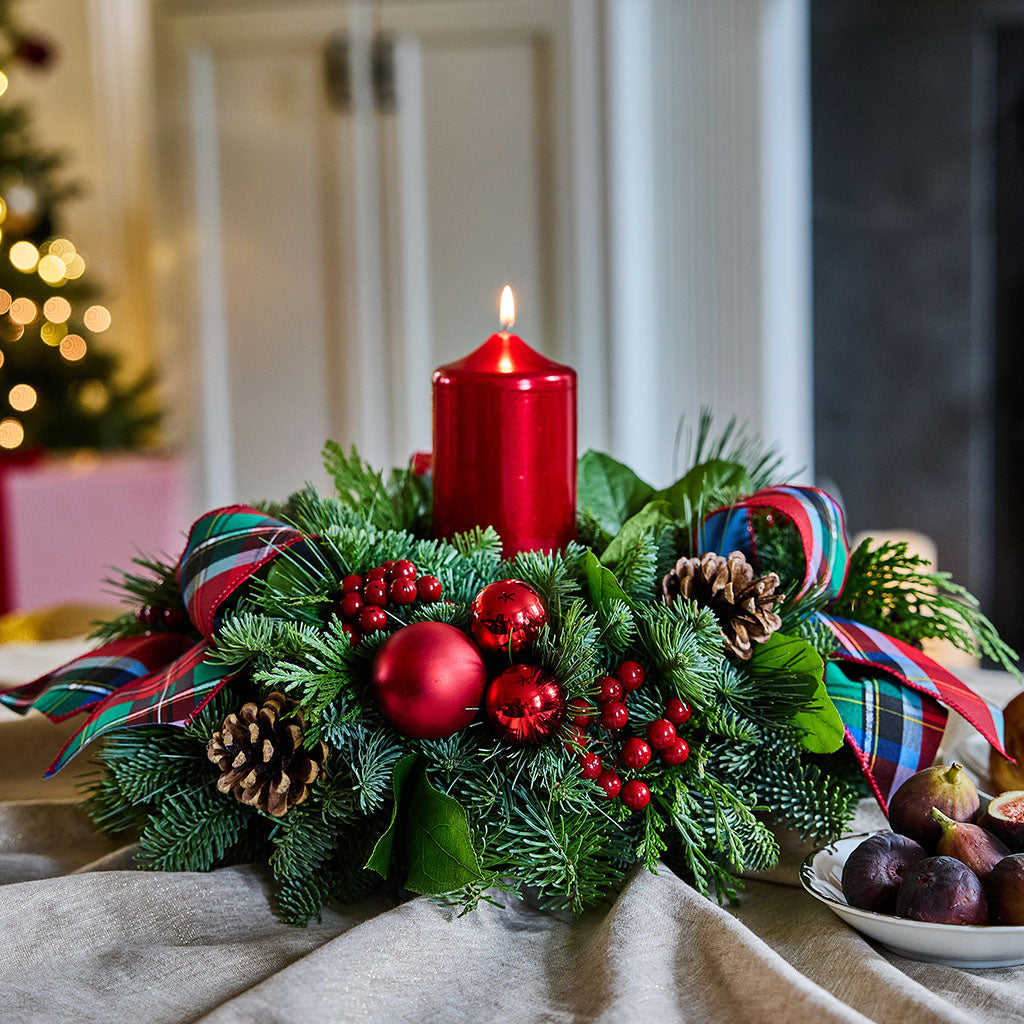 A festive Christmas wreath centerpiece on a draped tablecloth, featuring a lit red pillar candle, red ornaments, pinecones, red berries, and plaid ribbon bows, with a softly blurred holiday background.