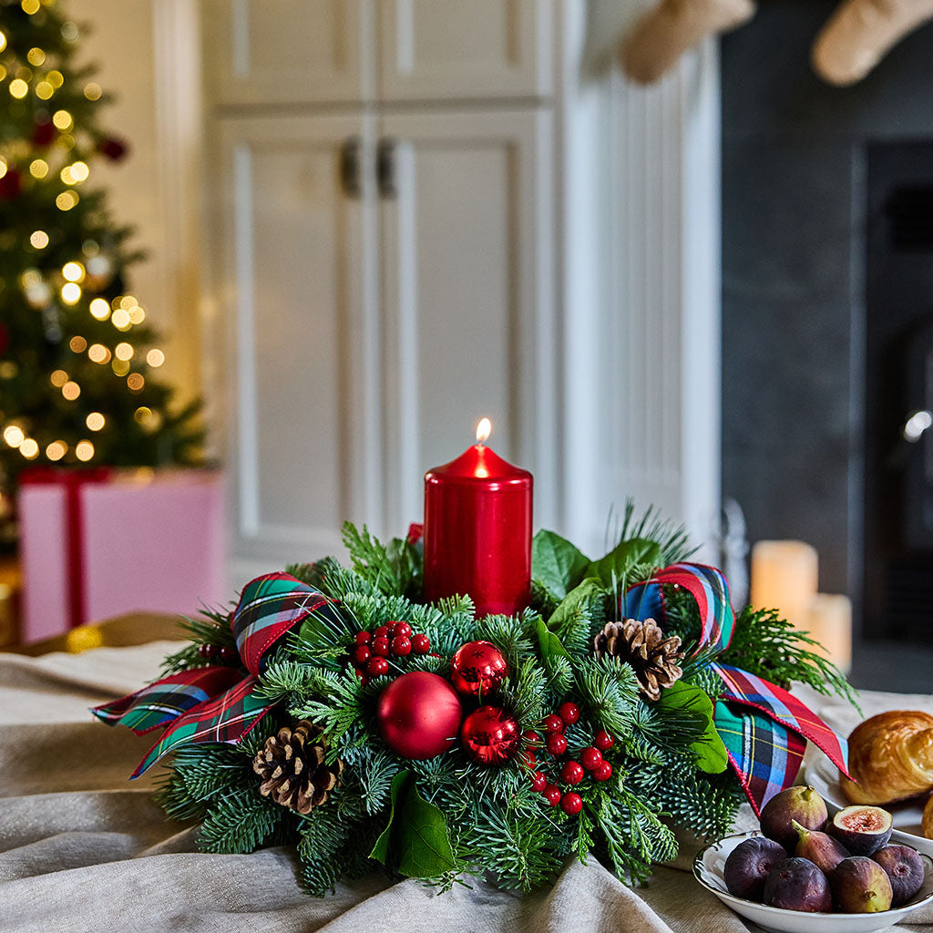A festive Christmas wreath centerpiece on a table, featuring a lit red pillar candle, green pine branches, red ornaments, pinecones, and plaid ribbon bows, with a blurred holiday home background.
