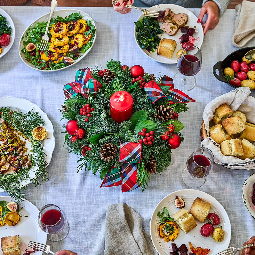 A festive holiday table centerpiece made of evergreen branches, red berries, pinecones, and red plaid ribbon bows, arranged on a white tablecloth with plates of food and people's hands visible around the table.