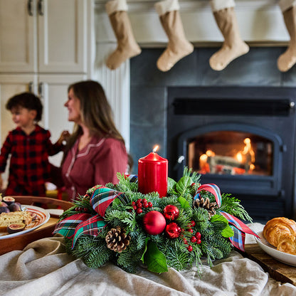 A festive Christmas wreath centerpiece with a lit red pillar candle, red ornaments, pinecones, and plaid ribbon, set on a table in a cozy living room with a fireplace and a woman and child blurred in the background.