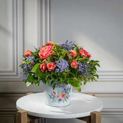 Fresh salal and green huckleberry with faux coral and peach roses, faux lavender Queen Anne’s Lace, and other faux purple flowers in a metal white floral container