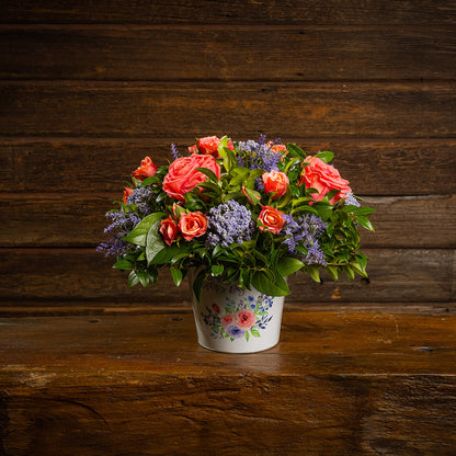 Fresh salal and green huckleberry with faux coral and peach roses, faux lavender Queen Anne’s Lace, and other faux purple flowers in a metal white floral container