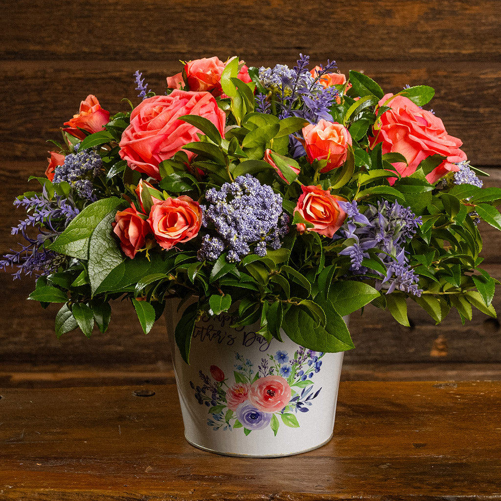 Fresh salal and green huckleberry with faux coral and peach roses, faux lavender Queen Anne’s Lace, and other faux purple flowers in a metal white floral container