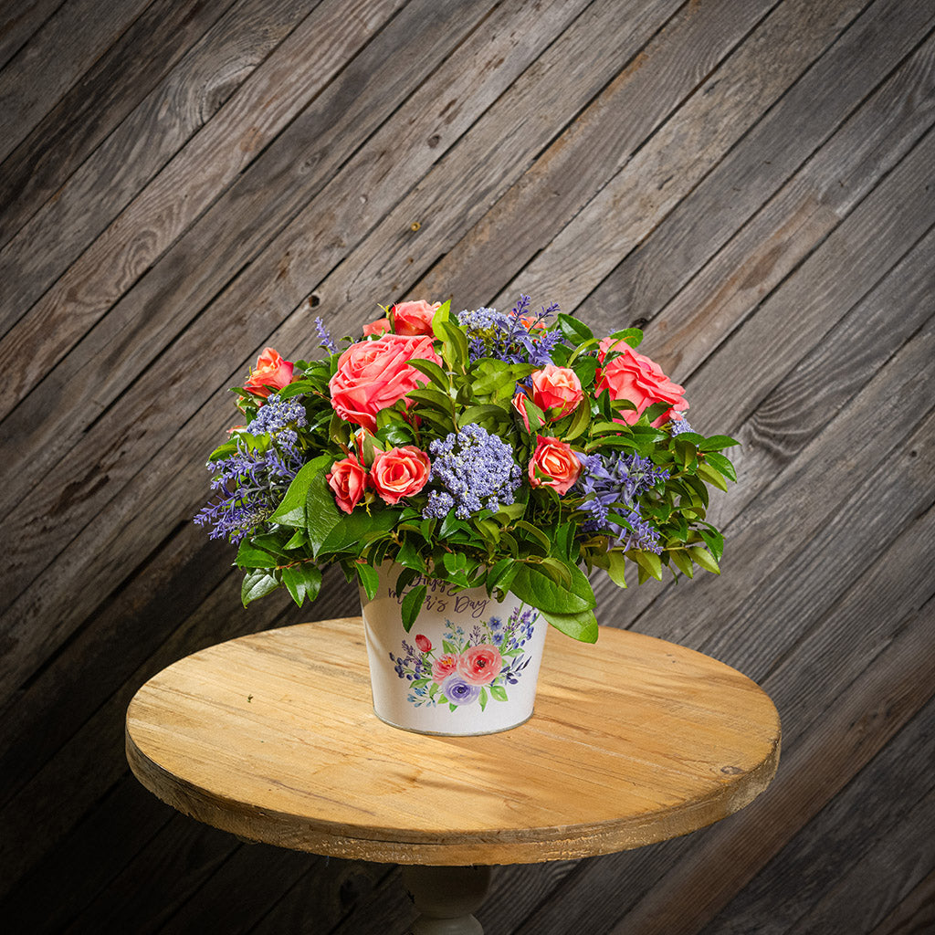 Fresh salal and green huckleberry with faux coral and peach roses, faux lavender Queen Anne’s Lace, and other faux purple flowers in a metal white floral container