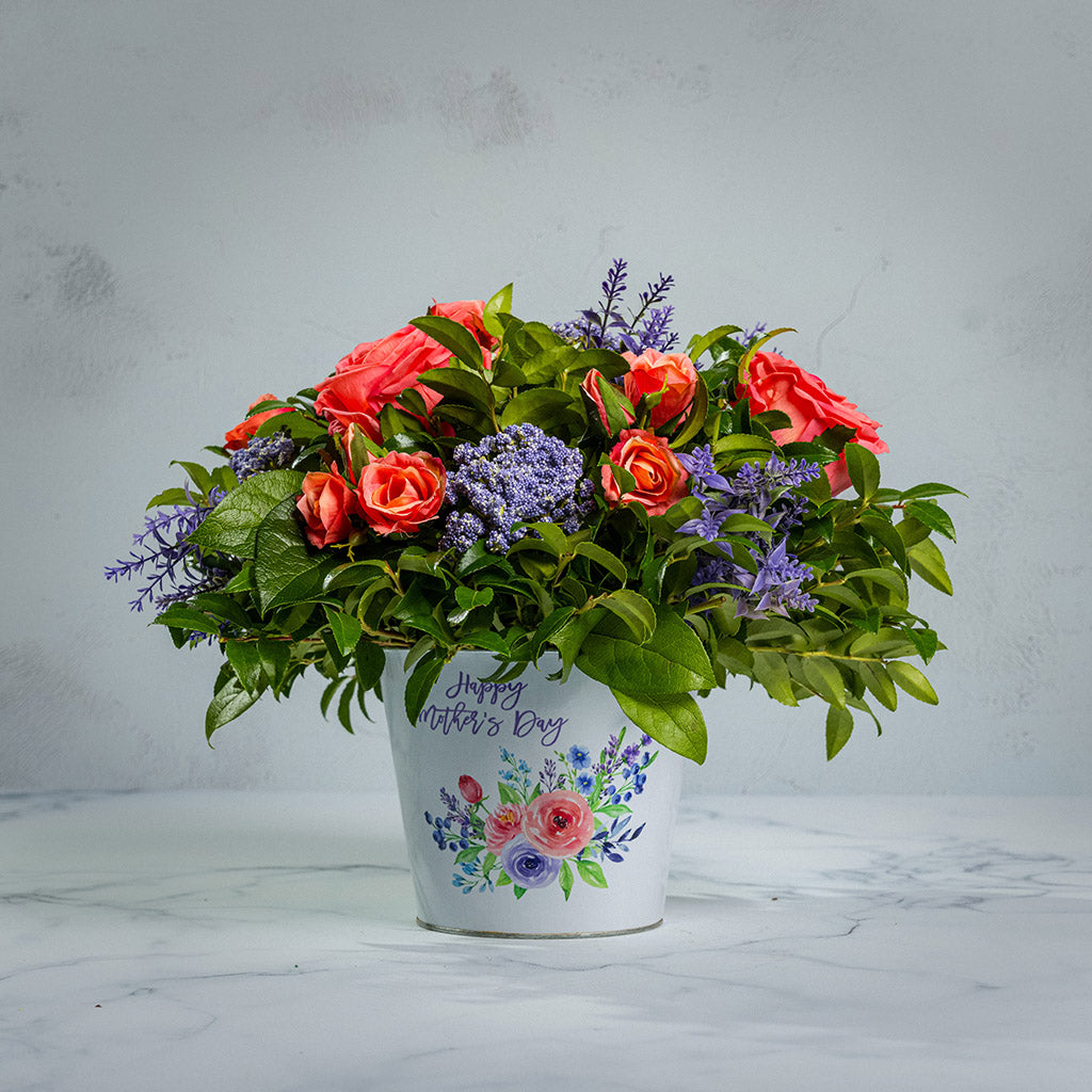 Fresh salal and green huckleberry with faux coral and peach roses, faux lavender Queen Anne’s Lace, and other faux purple flowers in a metal white floral container