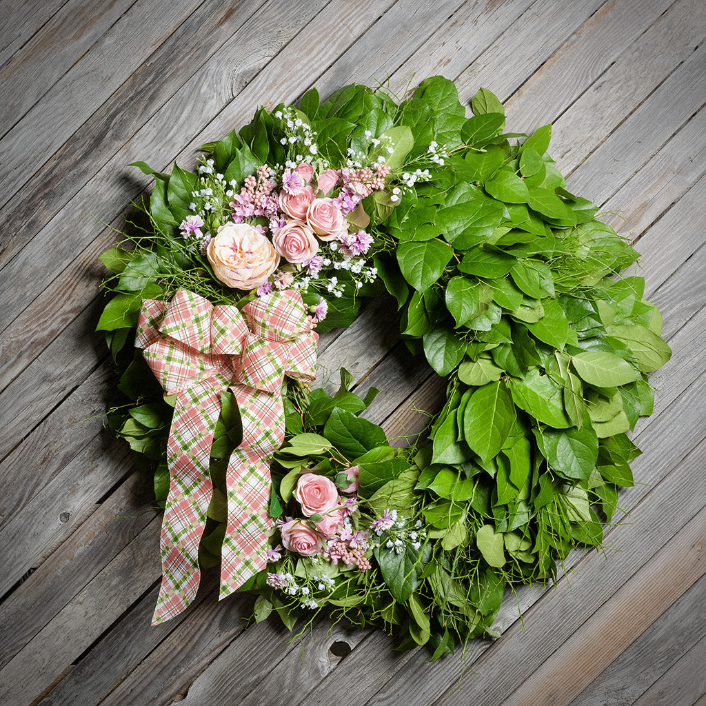 Green leaf wreath with pink flowers and a plaid ribbon on a wooden surface