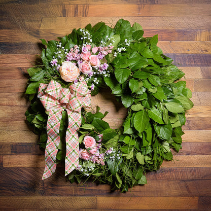 Green leaf wreath with pink flowers and a plaid bow on a wooden surface