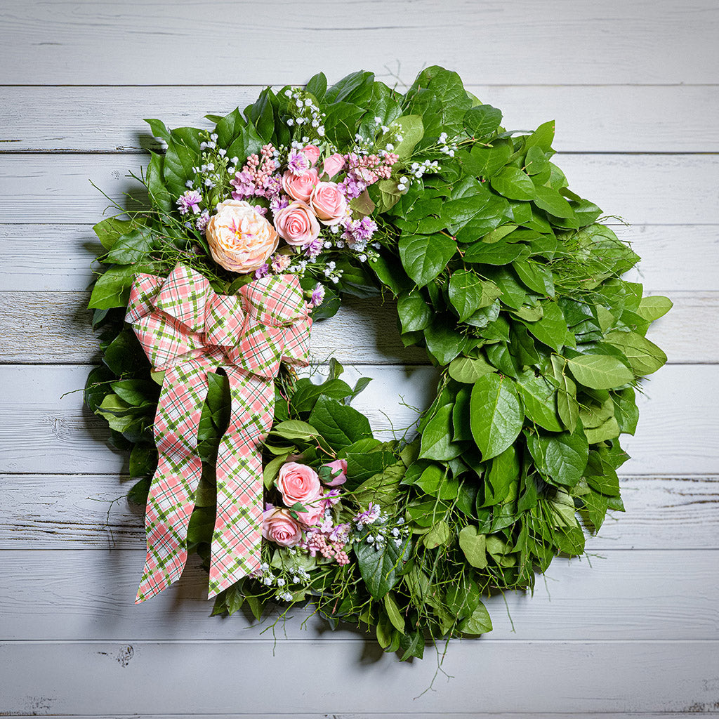 Green leaf wreath with pink flowers and a plaid bow on a light wooden background