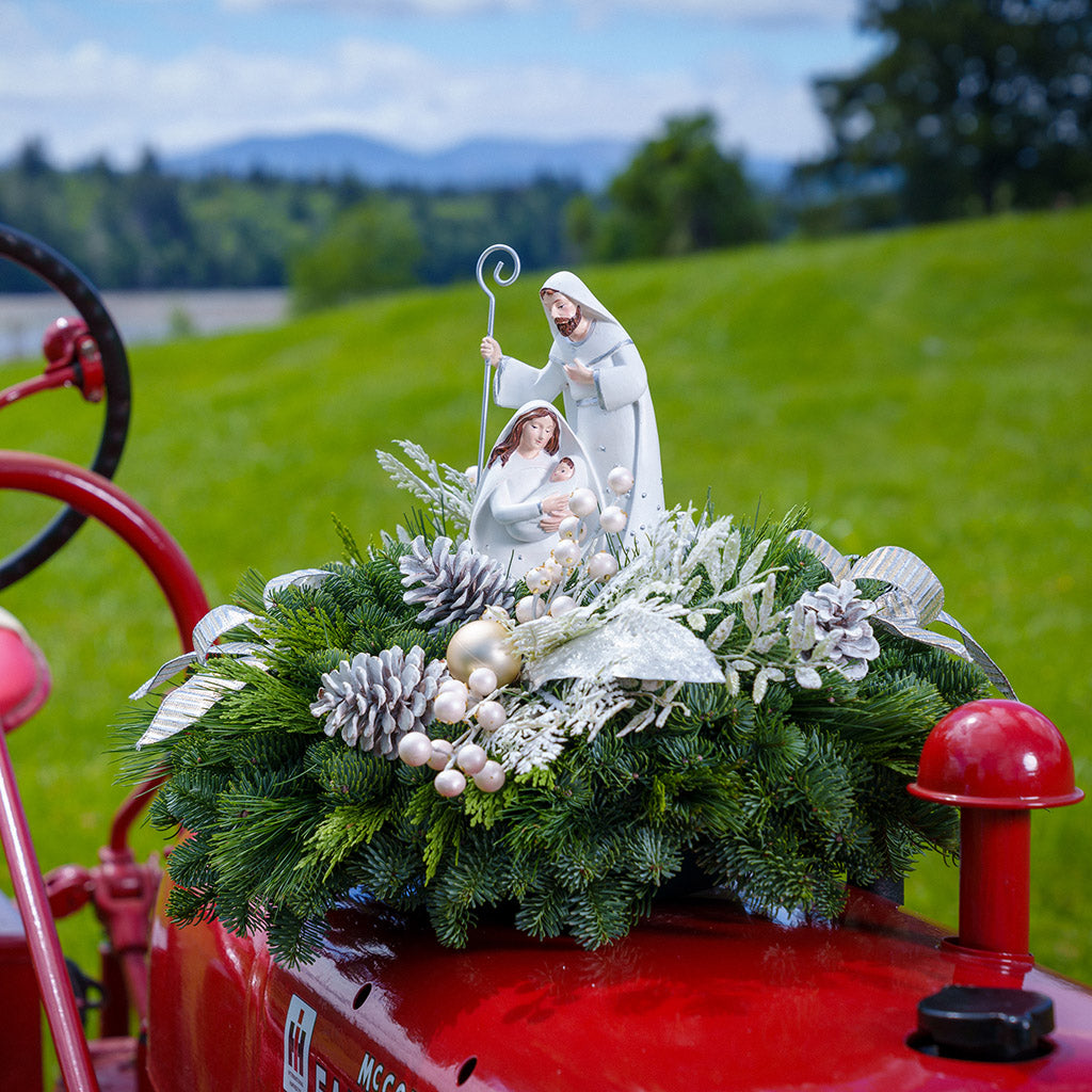 An evergreen arrangement of noble fir, cedar, and pine with frosted pinecones, champagne ball ornaments, white berries, glittery leaves, gold and silver bows, and a nativity scene decoration sitting on a red tractor with a green grass background.