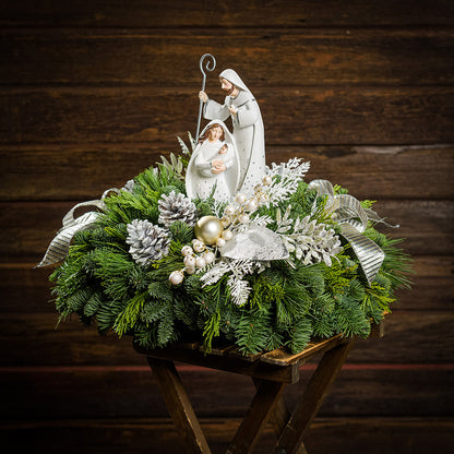 An evergreen arrangement of noble fir, cedar, and pine with frosted pinecones, champagne ball ornaments, white berries, glittery leaves, gold and silver bows, and a nativity scene decoration sitting on a table with a dark wood background.