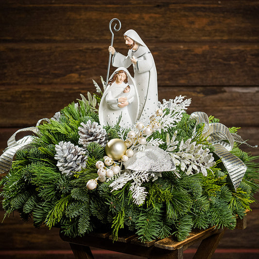 An evergreen arrangement of noble fir, cedar, and pine with frosted pinecones, champagne ball ornaments, white berries, glittery leaves, gold and silver bows, and a nativity scene decoration sitting on a table with a dark wood background.