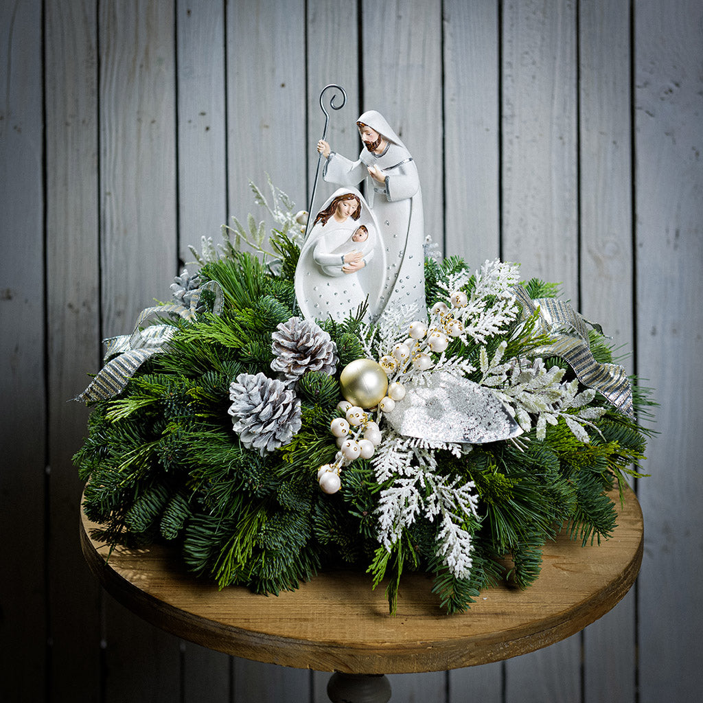 An evergreen arrangement of noble fir, cedar, and pine with frosted pinecones, champagne ball ornaments, white berries, glittery leaves, gold and silver bows, and a nativity scene decoration sitting on a table with a white wood background.