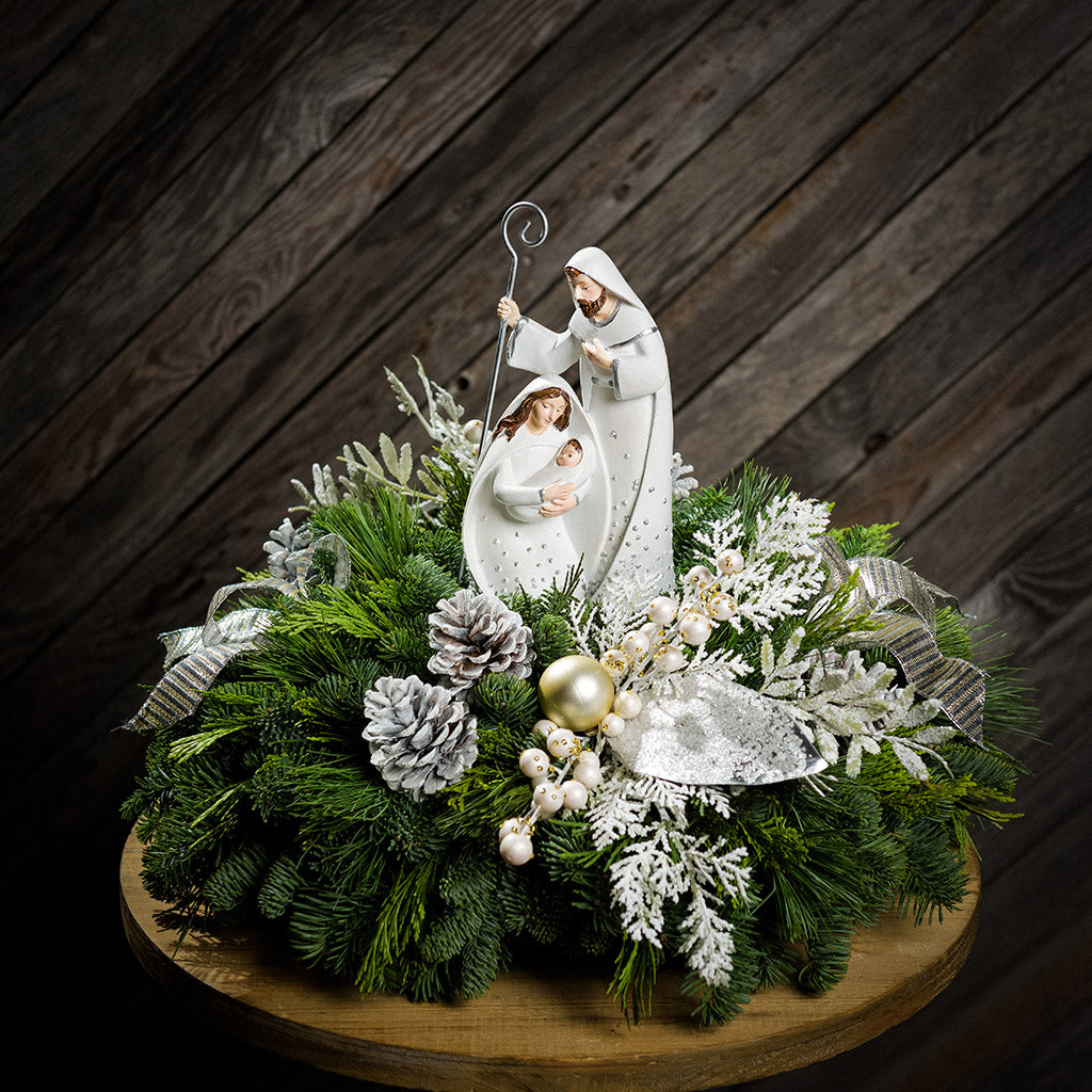 An evergreen arrangement of noble fir, cedar, and pine with frosted pinecones, champagne ball ornaments, white berries, glittery leaves, gold and silver bows, and a nativity scene decoration sitting on a table with a dark wood background.