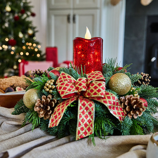 A festive Christmas wreath centerpiece with a red glitter candle, green pine branches, gold ornaments, pinecones, and a red-and-gold patterned bow, set on a beige fabric surface with a blurred holiday background.
