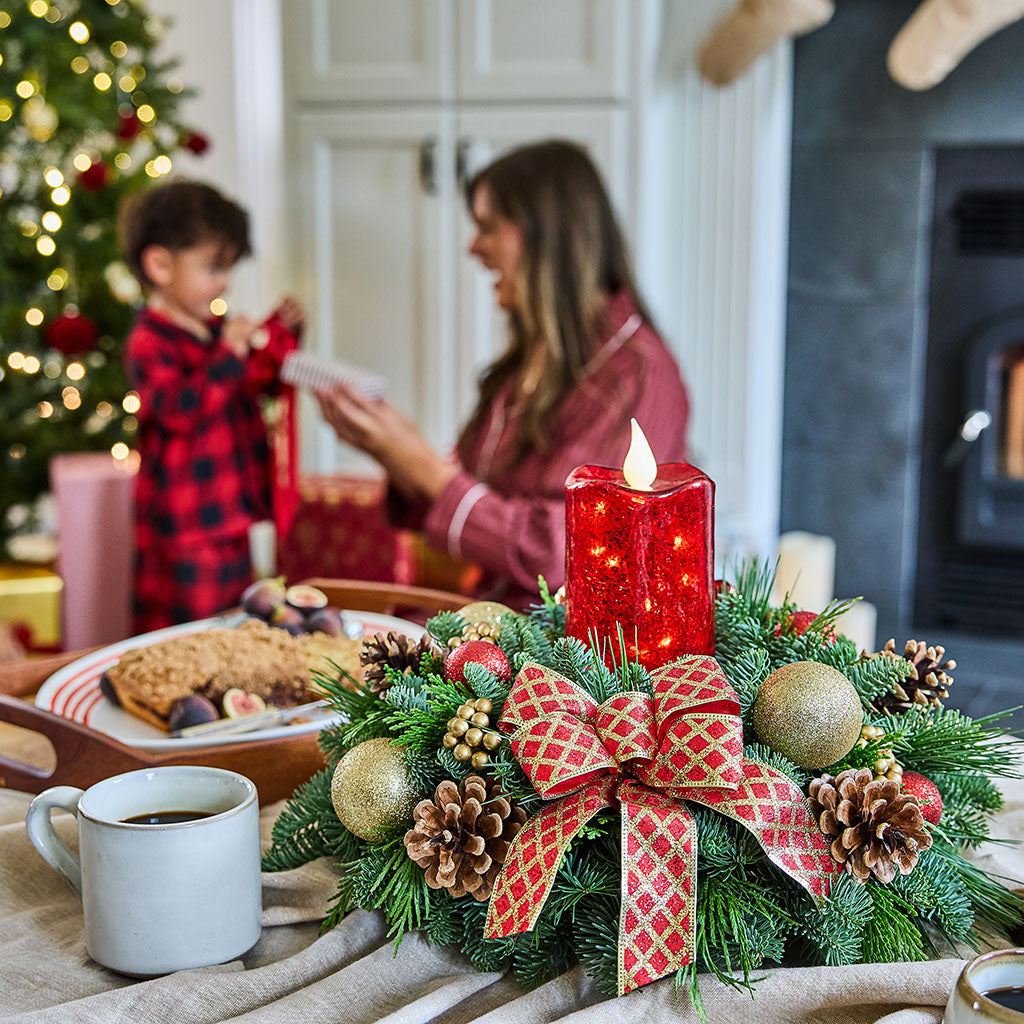 A festive Christmas wreath centerpiece on a table, featuring green pine branches, gold ornaments, pinecones, a red patterned ribbon bow, and a glowing red pillar candle. In the softly blurred background, a woman and child are gathered near a Christmas tree and fireplace in a cozy living room.