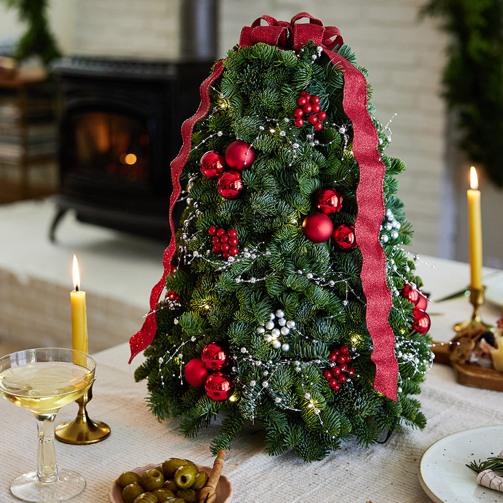 Tabletop tree of noble fir, red ball clusters, red berry clusters, silver berry clusters, a strand of metallic silver pearl garland and a red glittery ribbon close up