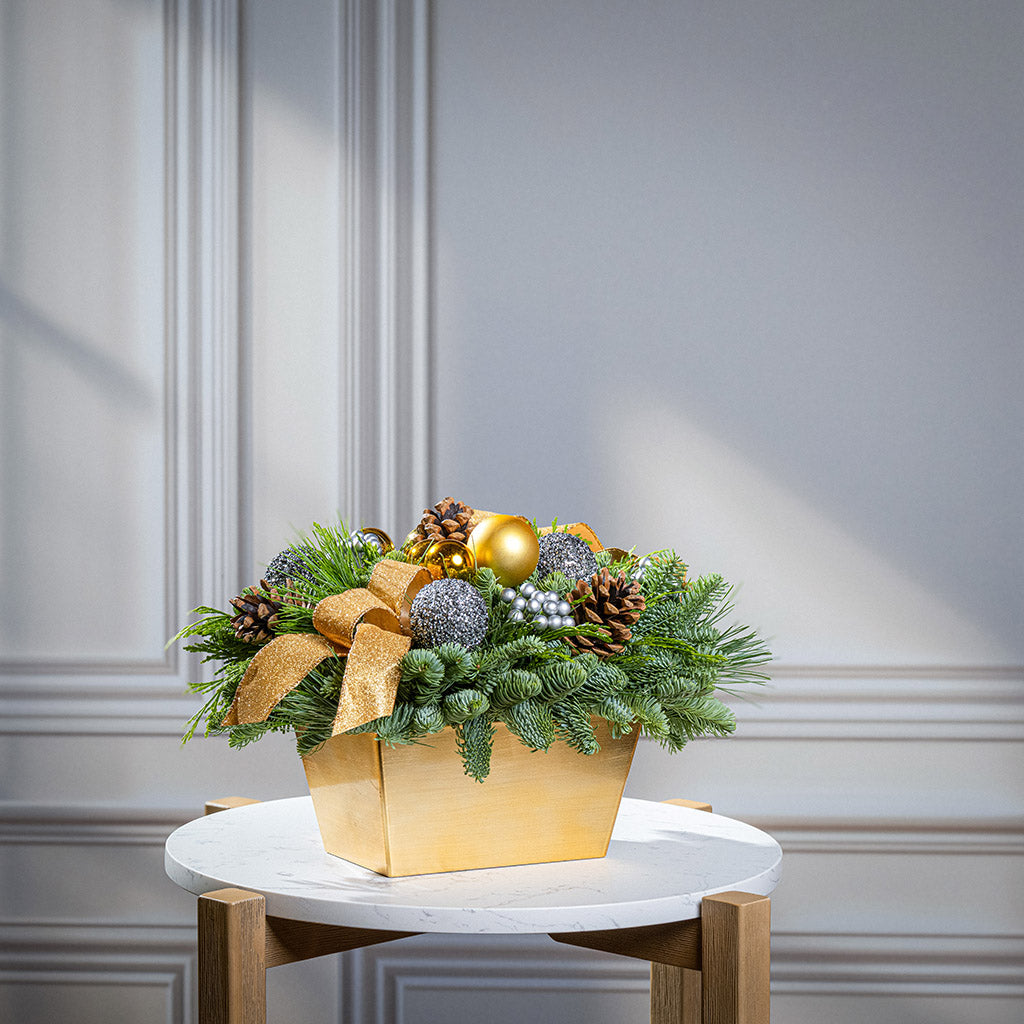 Decorative Christmas arrangement with greenery, ornaments, and a gold bow on a marble table.