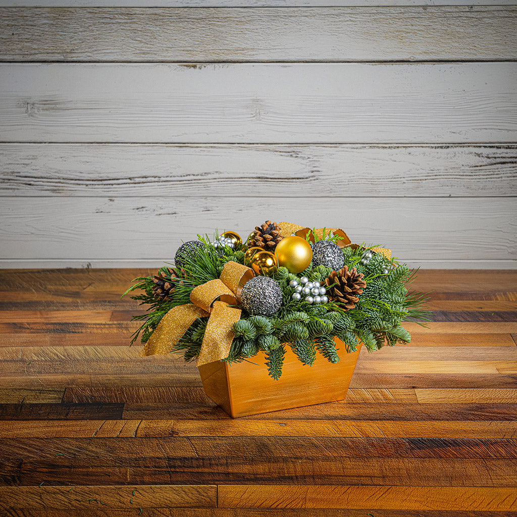 Decorative Christmas arrangement with greenery, ornaments, and a gold bow on a wooden surface.