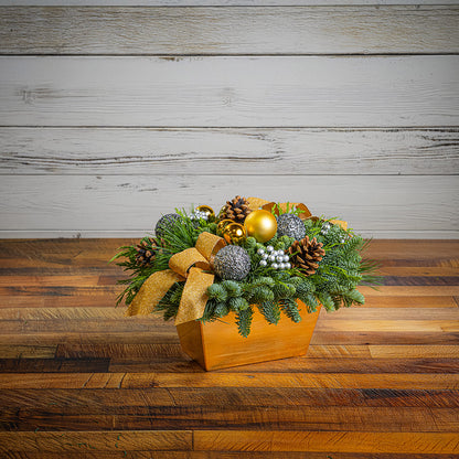 Decorative Christmas arrangement with greenery, ornaments, and a gold bow on a wooden surface.