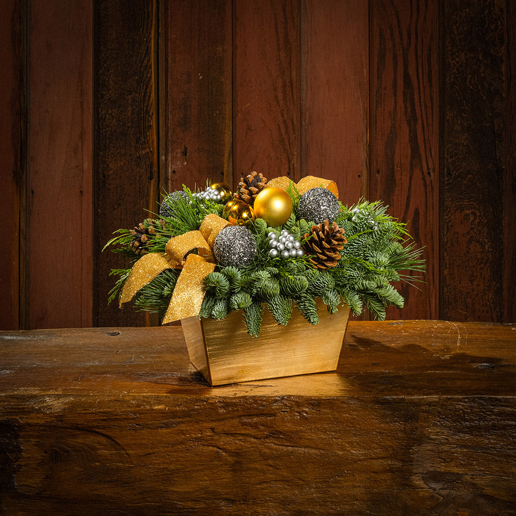 Decorative Christmas arrangement with gold bow, pine cones, and ornaments on a wooden surface.