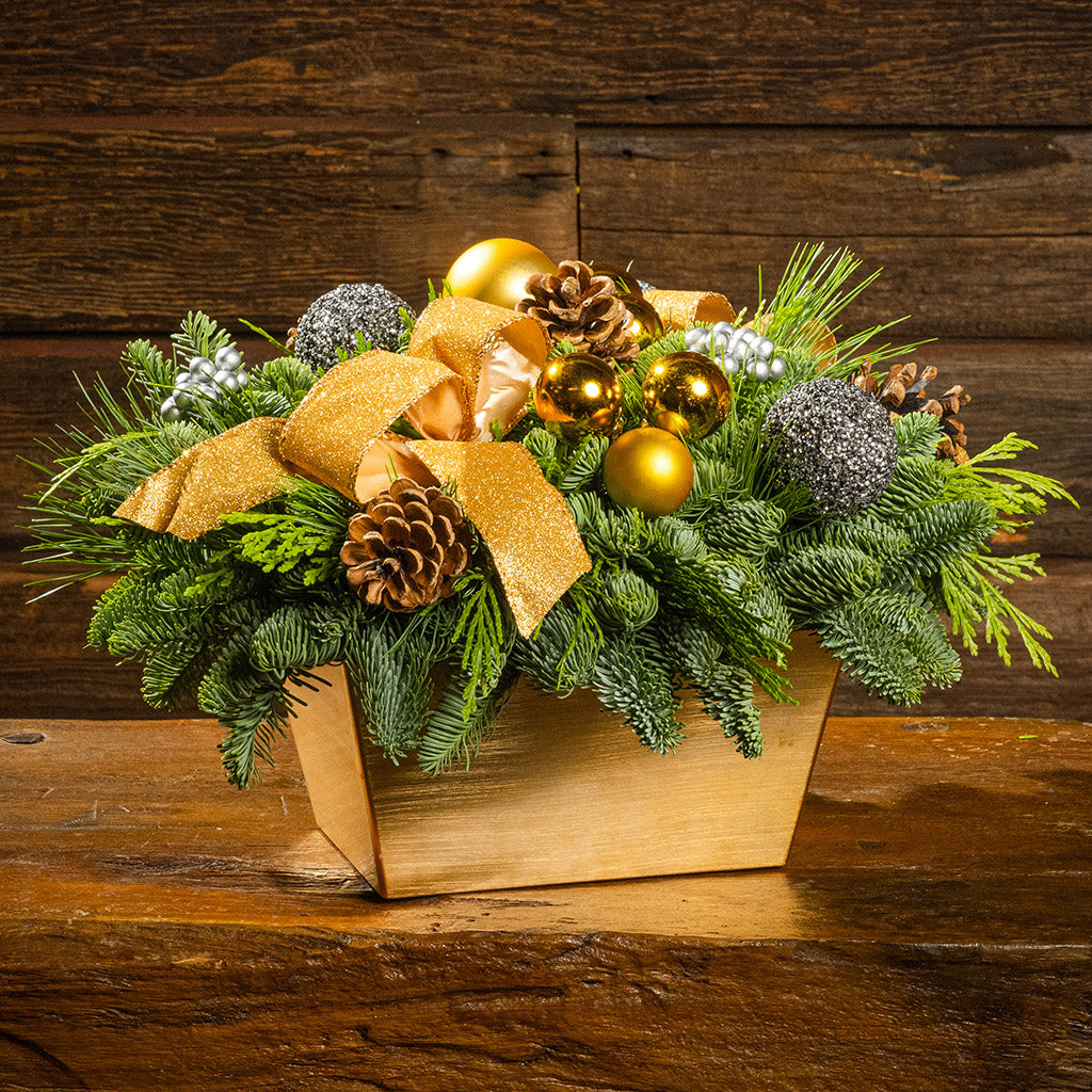 Decorative Christmas arrangement with greenery, gold ornaments, and a bow on a wooden surface.