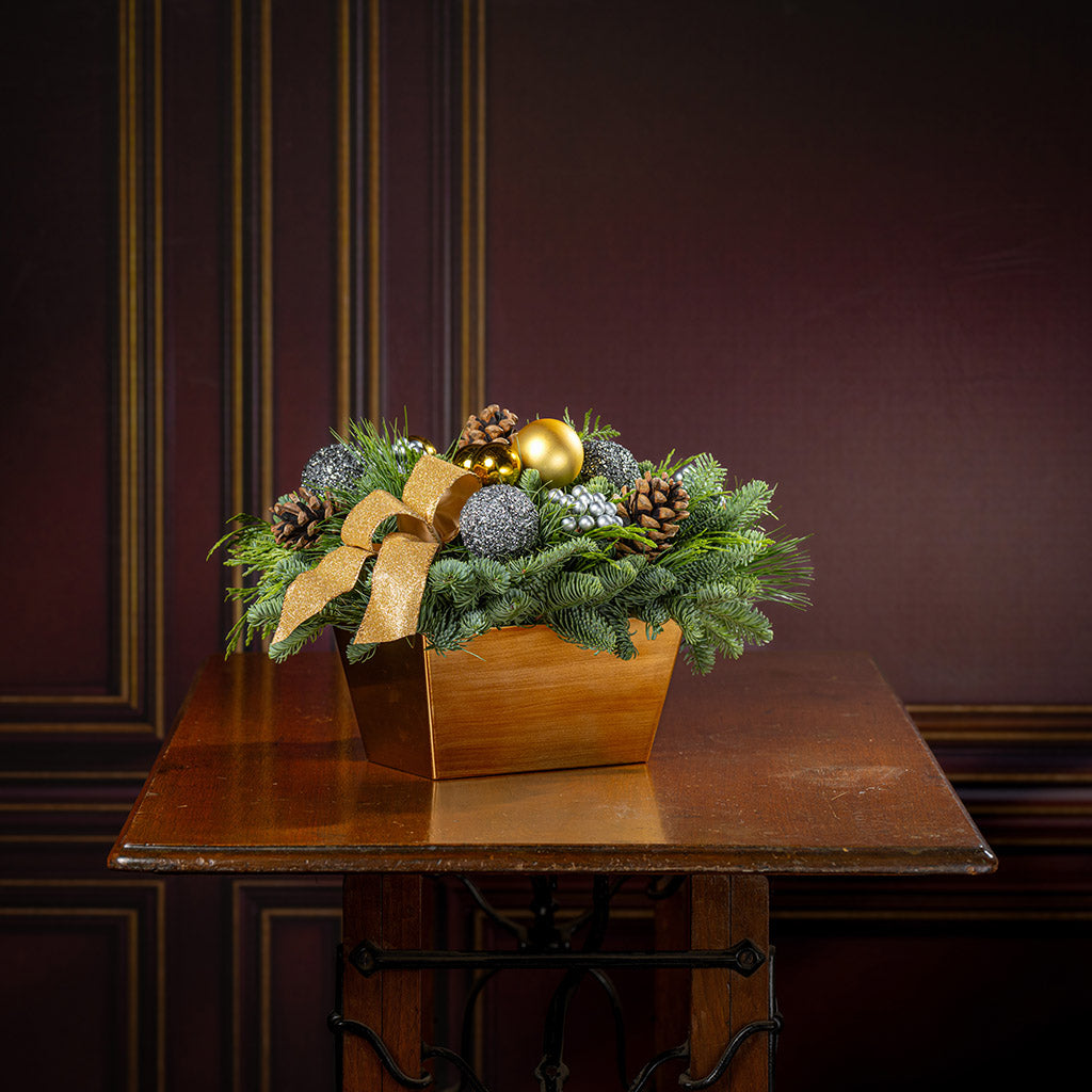 Decorative Christmas arrangement with greenery, ornaments, and a gold bow on a wooden table.