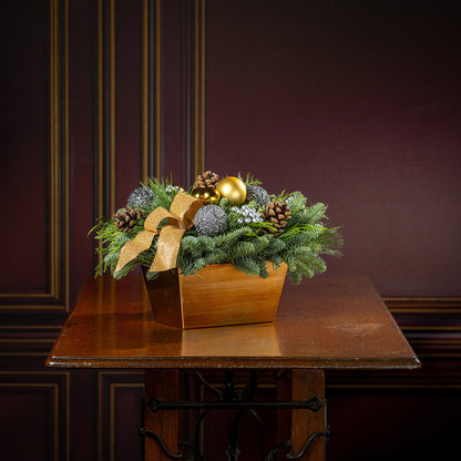 Decorative Christmas arrangement with greenery, ornaments, and a gold bow on a wooden table.