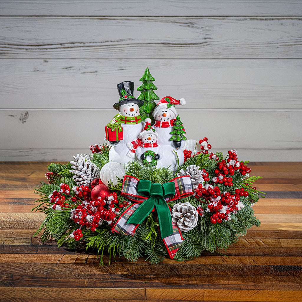 A festive Christmas wreath centerpiece featuring three stacked snowman figurines, red berries, pinecones, green pine branches, a red-and-white plaid bow, and a small green tree, displayed on a wooden surface against a white wood plank wall.