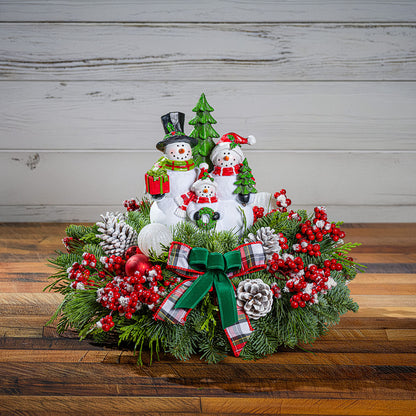 A festive Christmas wreath centerpiece featuring three stacked snowman figurines, red berries, pinecones, green pine branches, a red-and-white plaid bow, and a small green tree, displayed on a wooden surface against a white wood plank wall.