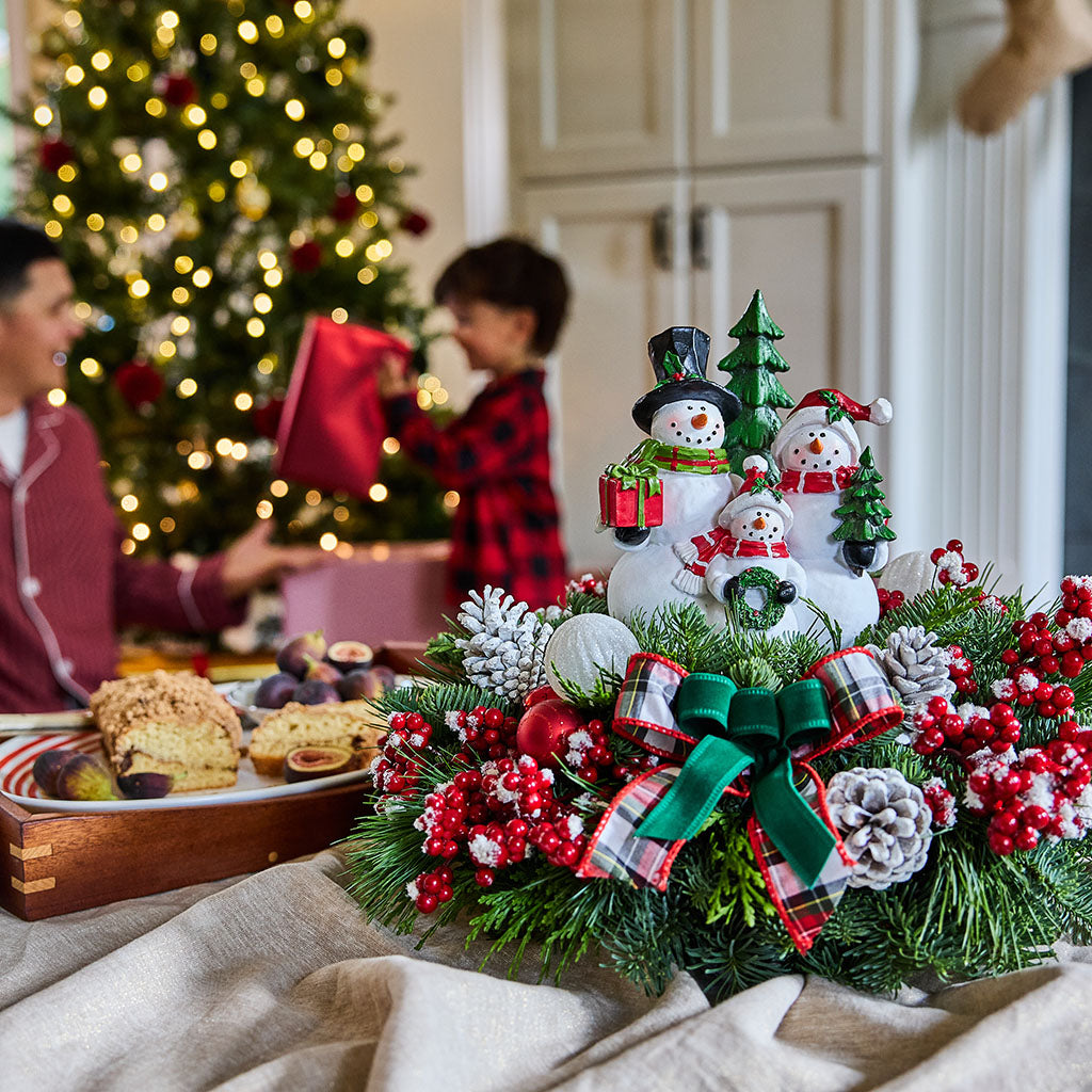 A festive Christmas snowman figurine centerpiece displayed on a table in a cozy holiday home setting, with a blurred Christmas tree and children in the background.