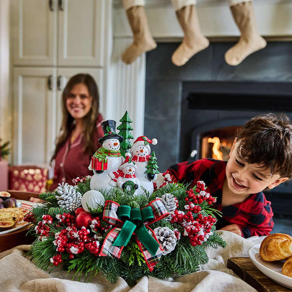A festive Christmas centerpiece with snowmen, pinecones, red berries, and a green ribbon bow sits on a table in a cozy living room setting, with a child in a red plaid shirt smiling in the foreground and a woman blurred in the background near a fireplace.