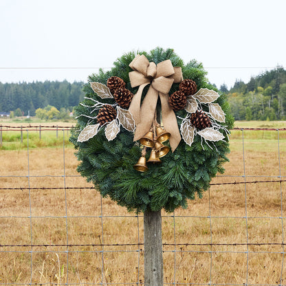 A green holiday wreath with pinecones, white leaf accents, a large tan burlap bow, and gold bells, hanging on a wooden post in a rural field with a wire fence and trees in the background.