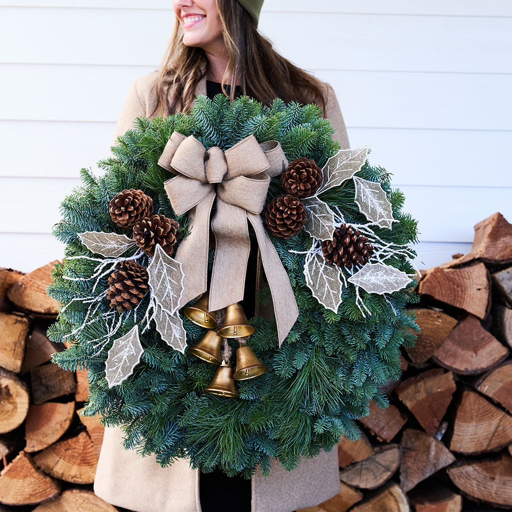 A woman holds a large green Christmas wreath decorated with a beige burlap bow, brown pinecones, white frosted leaves, and gold bells, set against a background of stacked firewood and a white paneled wall.