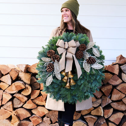 A woman wearing an olive green beanie and a beige coat holds a large green Christmas wreath decorated with a beige ribbon bow, pinecones, white leaf ornaments, and small gold bells, standing in front of a stacked woodpile against a white siding wall.