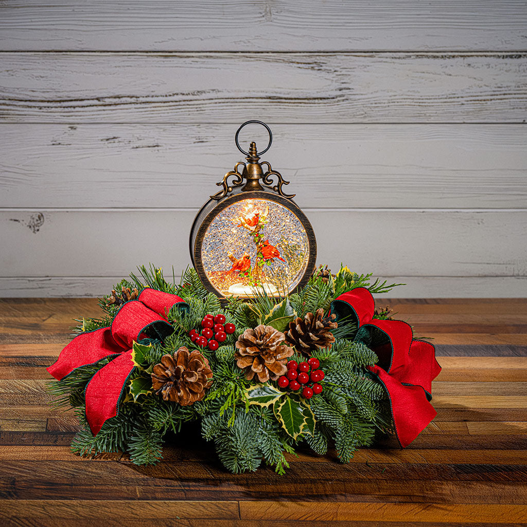 A festive Christmas wreath centerpiece with a hanging lantern ornament, green pine branches, red fabric bows, pinecones, and red berries, displayed on a wooden surface against a whitewashed wood plank background.