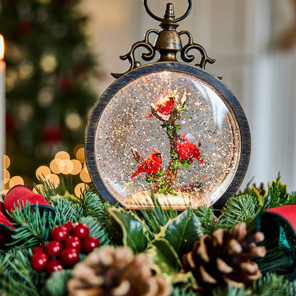 A round decorative snow globe ornament with a black rim and ornate metal top, filled with glitter and featuring two red birds perched on a branch. The ornament is displayed among holiday greenery, pinecones, and red berries in a softly blurred festive setting.