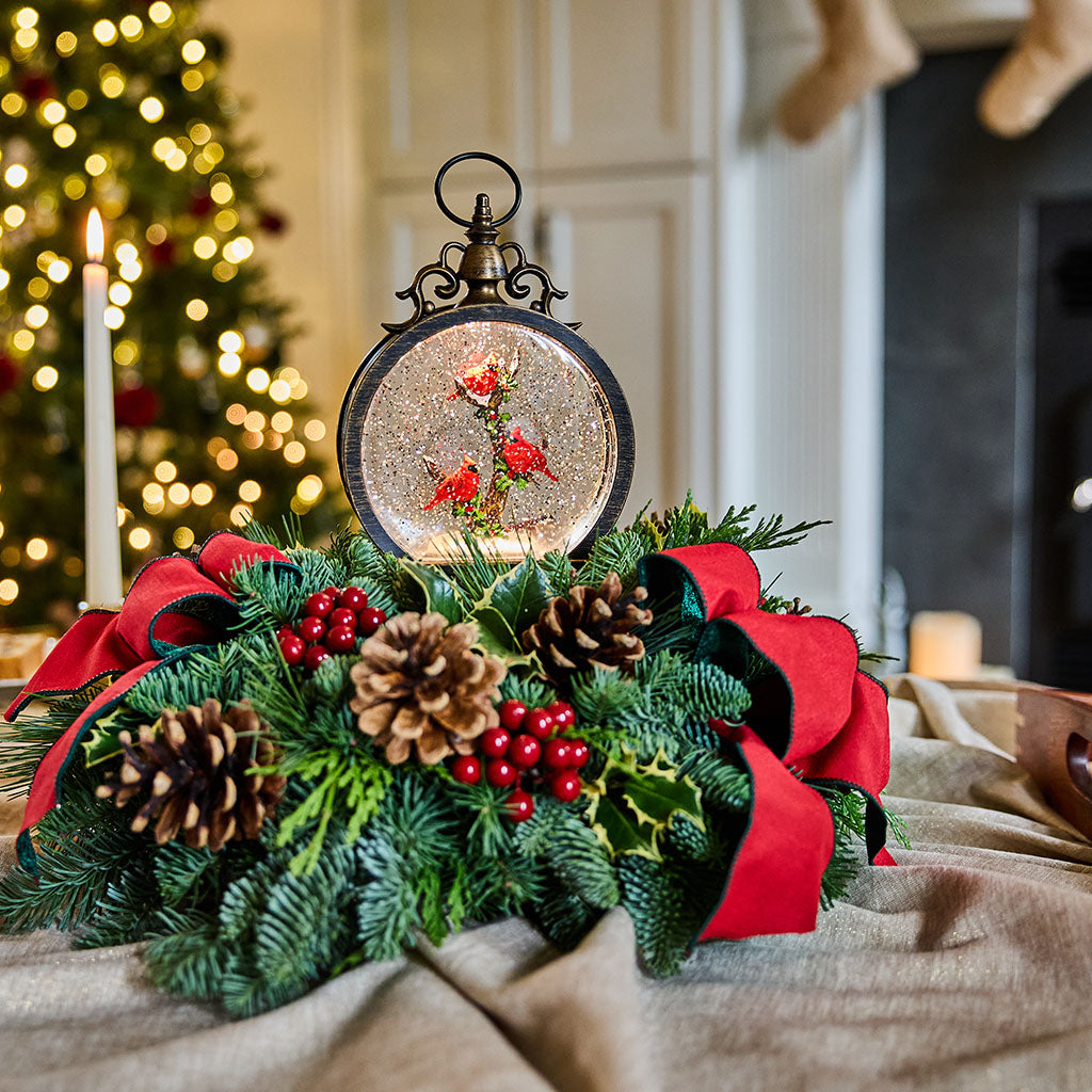 A festive Christmas wreath centerpiece with green pine branches, red ribbons, pinecones, red berries, and a round hanging ornament filled with glitter and small red floral details, displayed on a draped fabric surface with a softly blurred holiday interior in the background.
