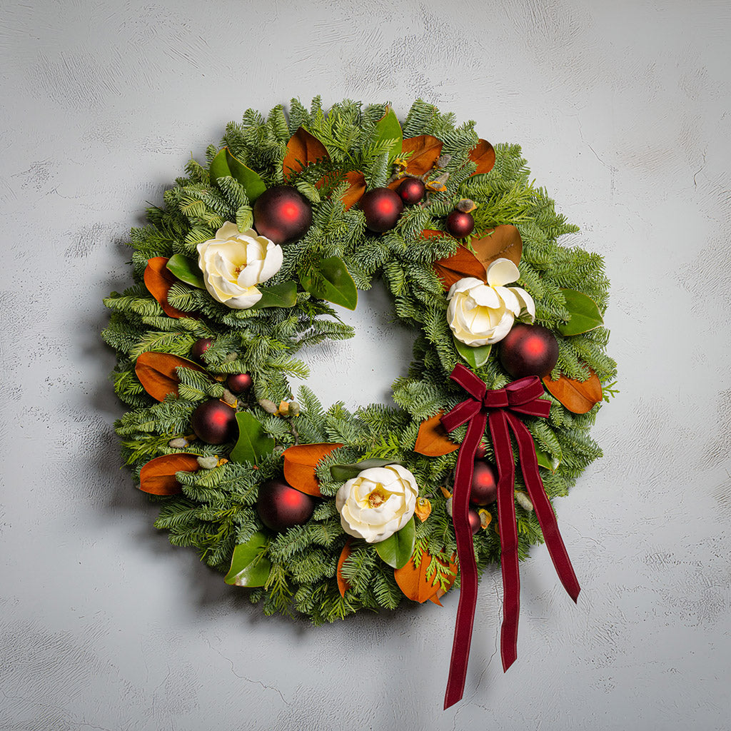 Decorative Christmas wreath with red ornaments, white flowers, and a red ribbon on a light gray background.