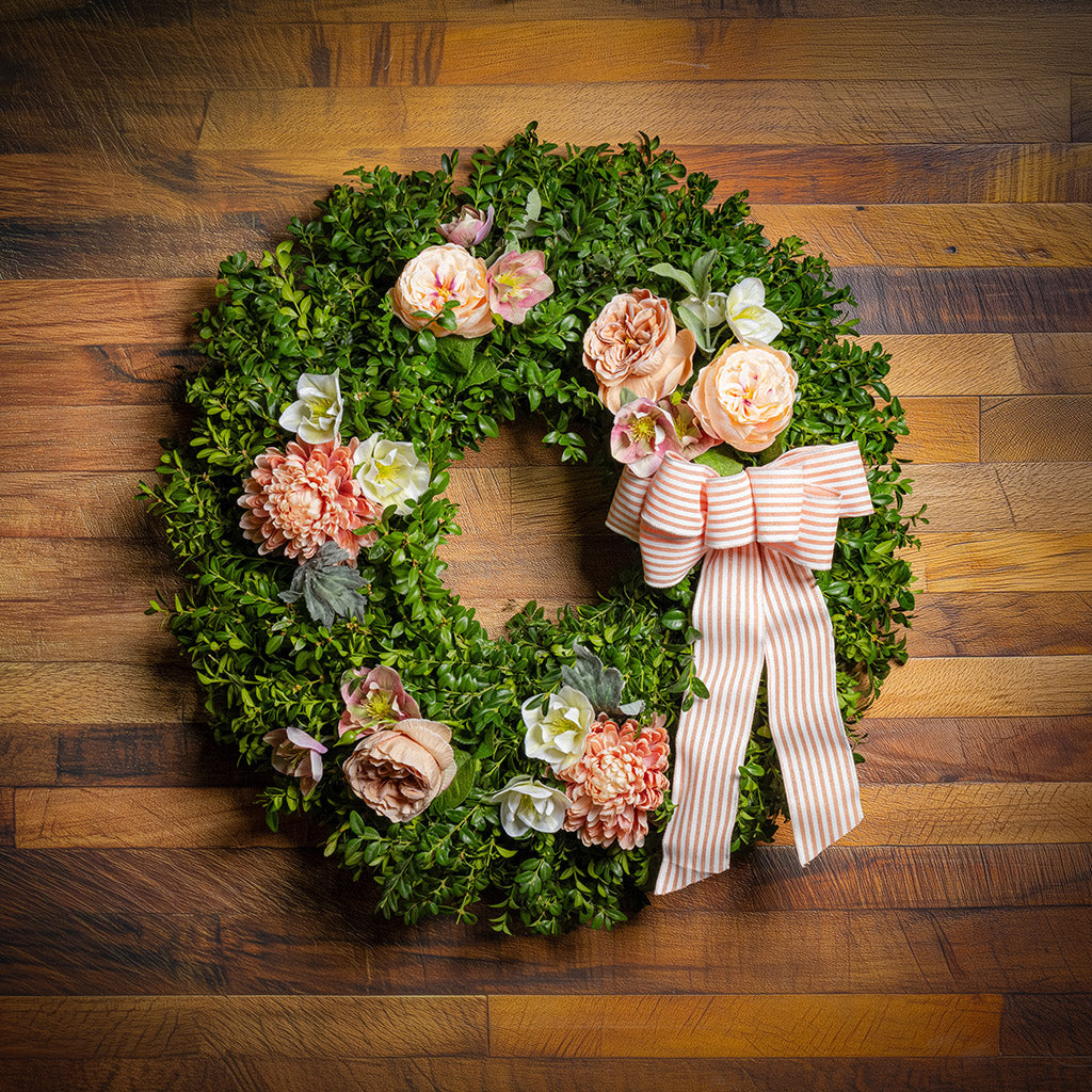 Fresh boxwood with faux peachy mums, faux peachy roses, and faux cream hellebore flowers with a peach and white striped bow