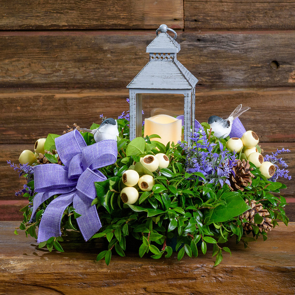 Decorative arrangement with a lantern, flowers, and a purple bow on a wooden surface.