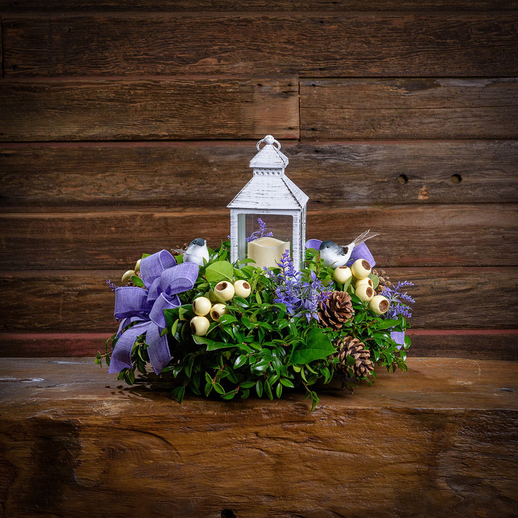Decorative arrangement with a lantern, flowers, and pinecones on a wooden surface.