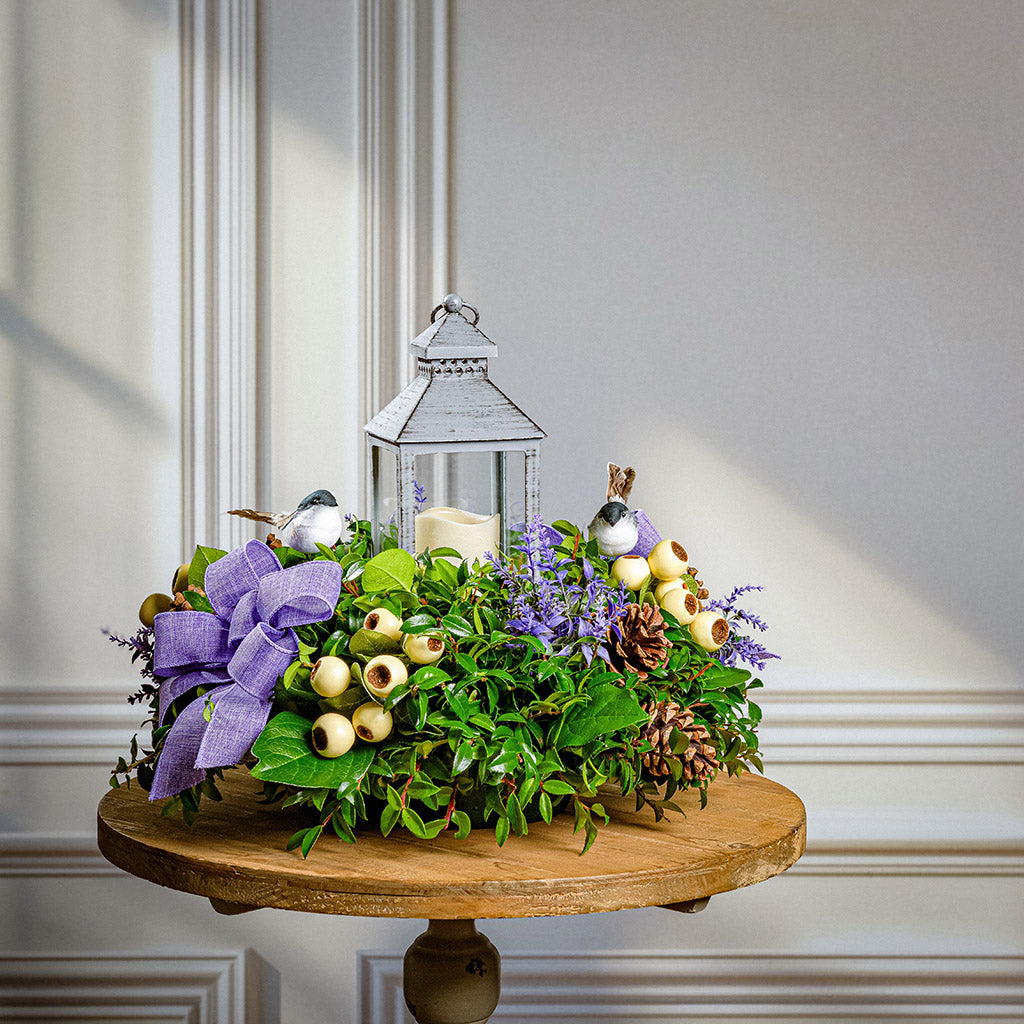 Decorative centerpiece with lantern, flowers, and candles on a wooden table.