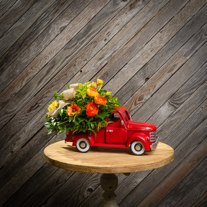 Fresh salal and green huckleberry with faux orange cosmos flowers, faux mini yellow roses, faux orange ranunculus flowers, and a natural linen bow in a red metal truck container