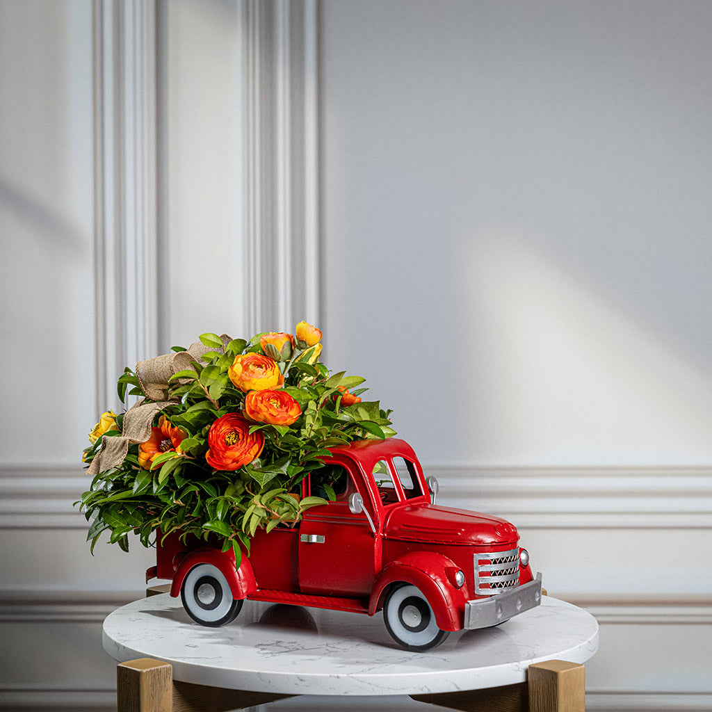 Fresh salal and green huckleberry with faux orange cosmos flowers, faux mini yellow roses, faux orange ranunculus flowers, and a natural linen bow in a red metal truck container